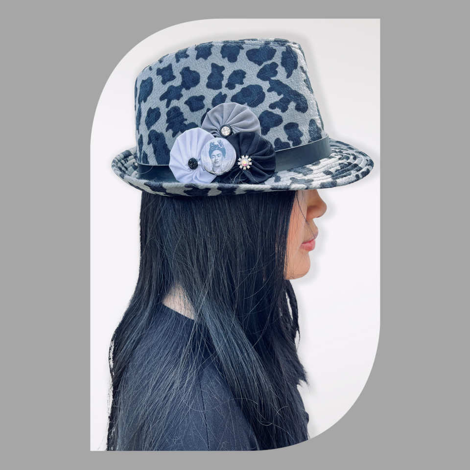 Woman facing right with black hair wearing a grey leopard hat adorned with grey and white fabric yo-yos, vintage  jewelry and a Frida Kahlo button