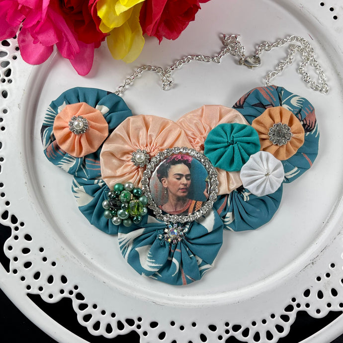 1940's Teal Fabric with Peach & White yo-yos & Teal Frida Pendant Necklace on a white plate with colorful flowers in the top left corner