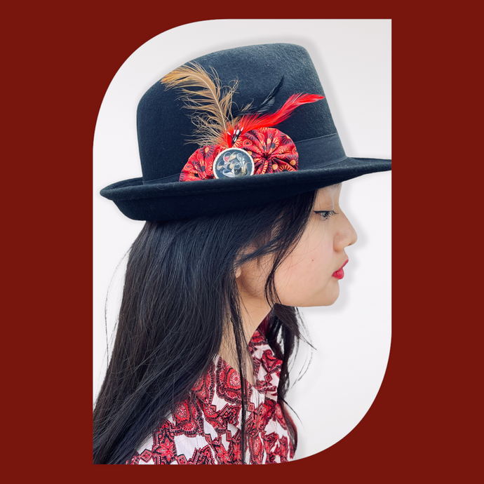 Black Fedora Hat with Feathers and Fabric Flowers on a model with long black hair staring right. The background is red